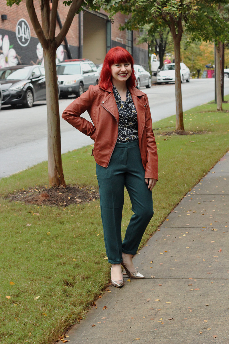 Smiling red-haired woman outdoors wearing a cognac-brown faux-leather moto biker jacket over a blue bow blouse with dark green skinny trousers and rose-gold pumps