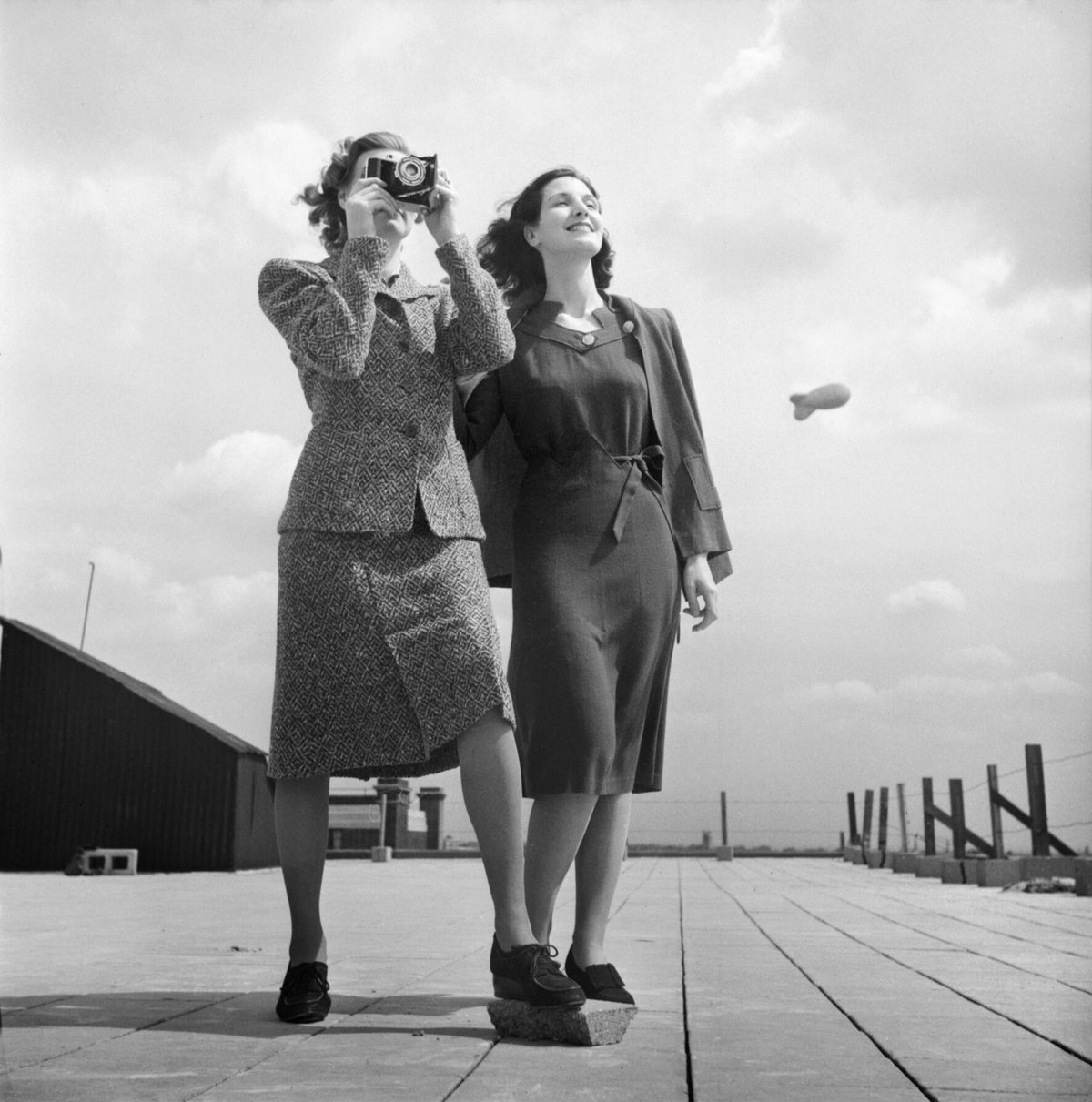 Two women posing on a London rooftop in 1943; the woman on the left holds a camera to her face and wears a patterned tweed jacket and matching skirt.