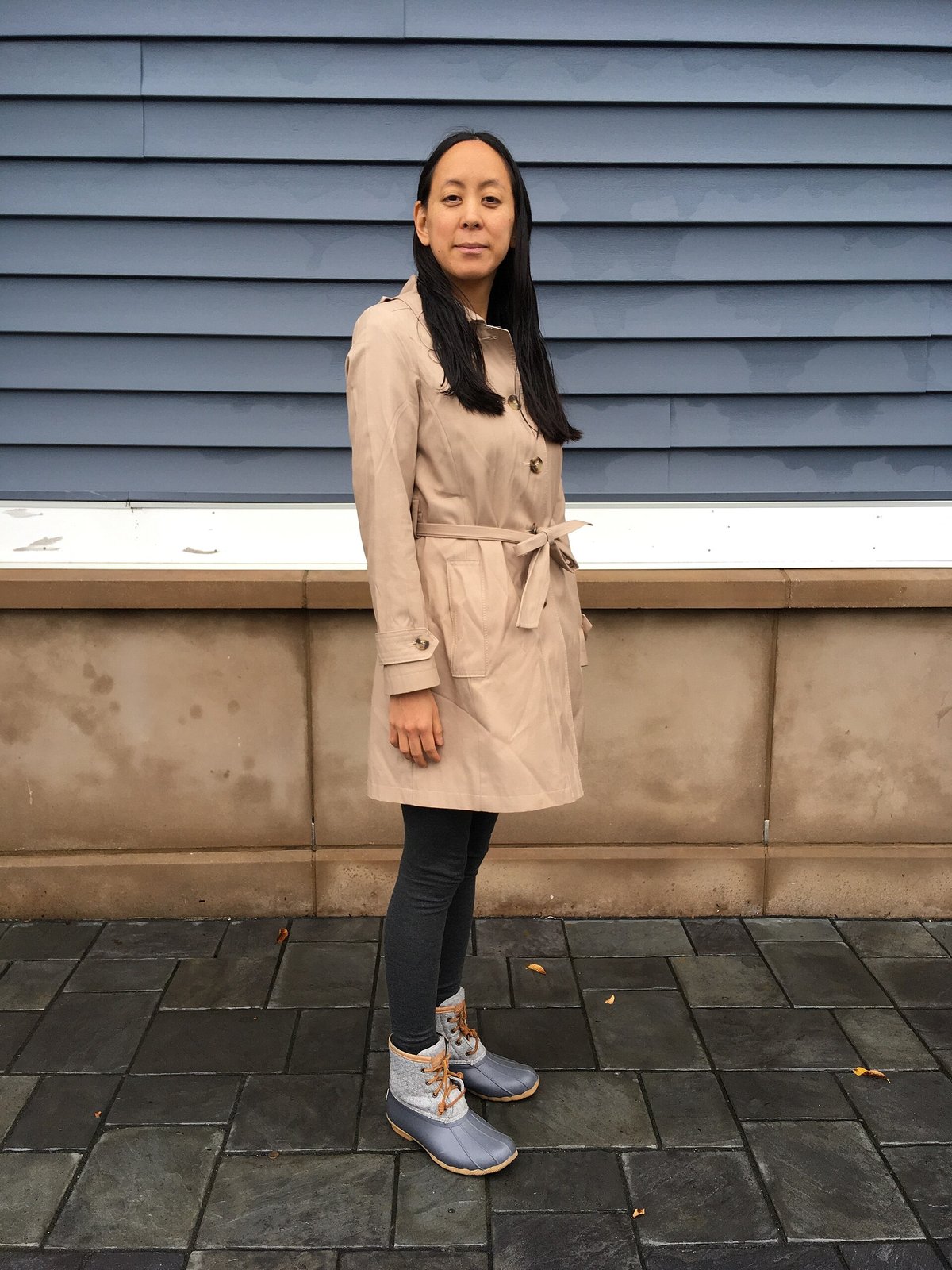 A woman standing against a blue wall wearing a beige belted trench coat over black leggings and grey lace-up duck boots