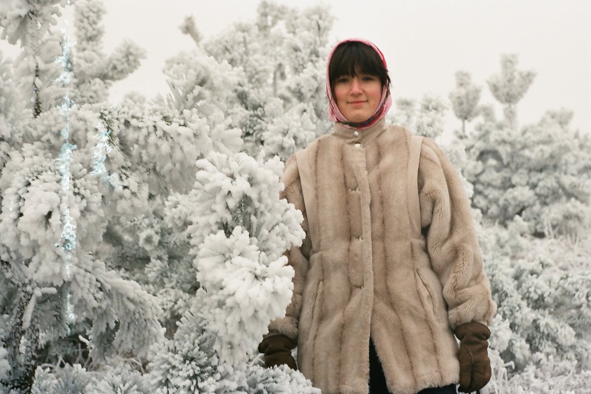 A woman among frost-covered trees wearing a beige fur coat with a stand-up collar, brown gloves and a pink headscarf.