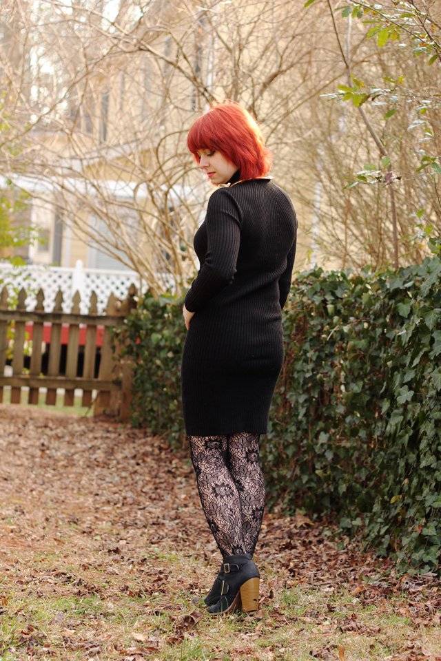 A red-haired woman outdoors wearing a black long-sleeved bodycon knit sweater dress with black lace tights and black ankle boots.