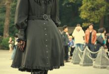 A woman with long auburn curly hair wearing a black gothic lolita dress with a ruffled collar, belt, lace tights and platform heels, posing outdoors