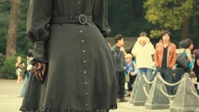 A woman with long auburn curly hair wearing a black gothic lolita dress with a ruffled collar, belt, lace tights and platform heels, posing outdoors