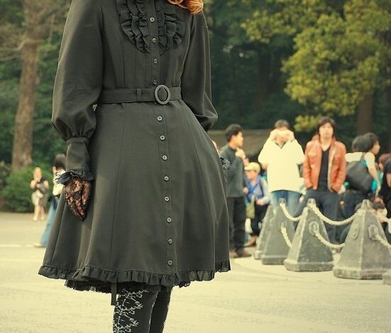 A woman with long auburn curly hair wearing a black gothic lolita dress with a ruffled collar, belt, lace tights and platform heels, posing outdoors
