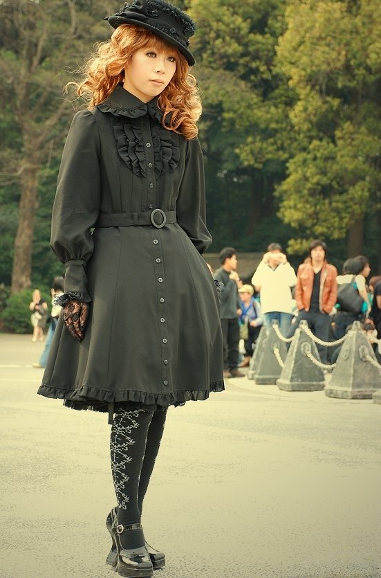 A woman with long auburn curly hair wearing a black gothic lolita dress with a ruffled collar, belt, lace tights and platform heels, posing outdoors