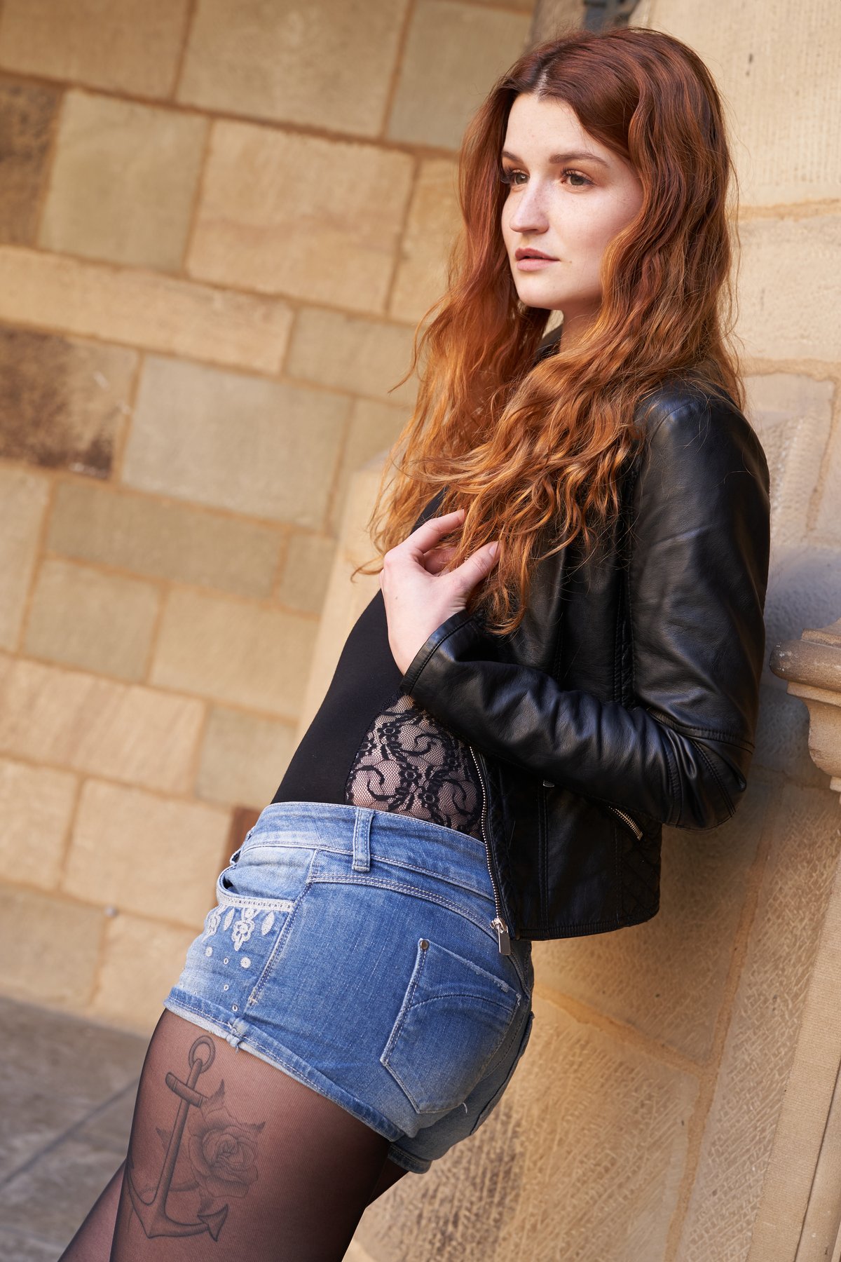 A woman with long wavy red hair wearing a black leather jacket open over a black lace-panel top, denim shorts and fishnet tights, leaning against a stone wall