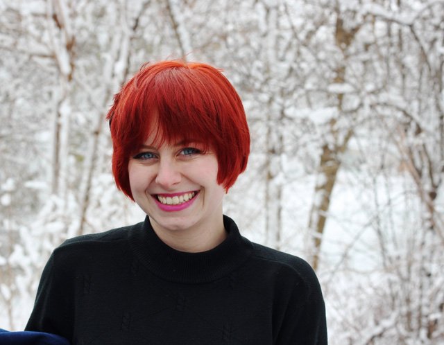 Smiling woman with a short red pixie haircut wearing a black mock-neck knit turtleneck sweater, standing outdoors in front of snow-covered trees.