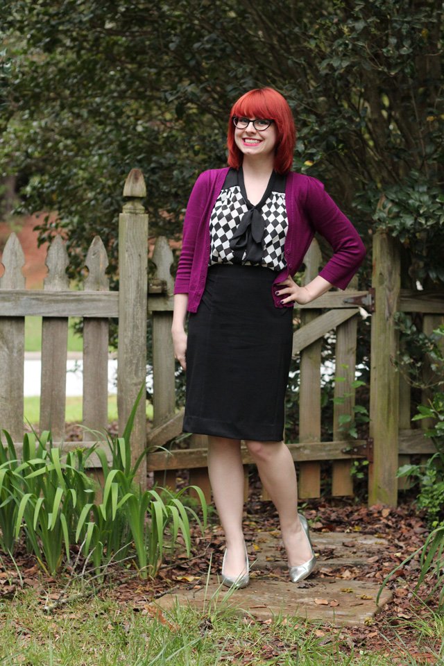 Woman with red hair standing outdoors wearing a black pencil skirt, a checkerboard bow blouse, a purple cardigan and silver pointed heels