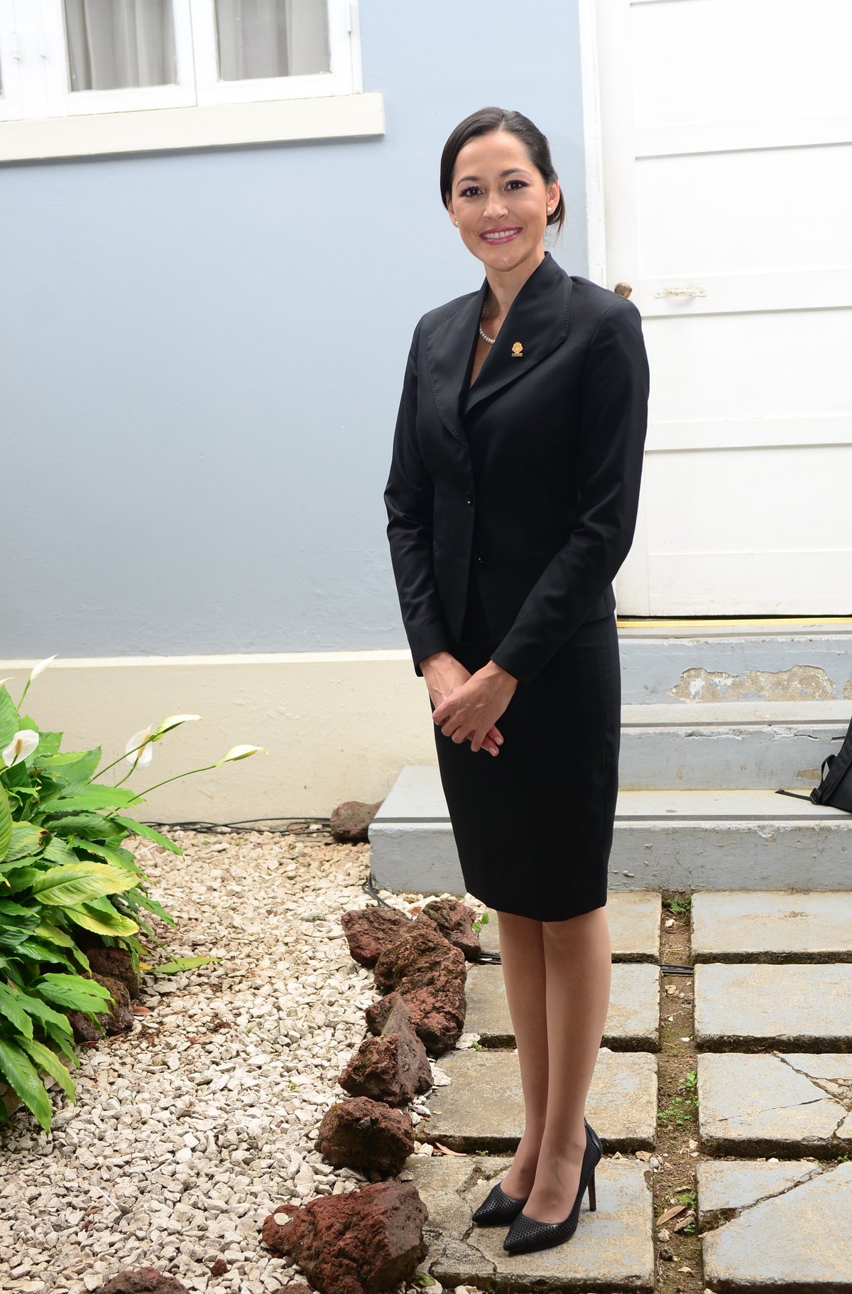Woman standing outdoors in a black skirt suit with a pearl necklace and black pointed heels