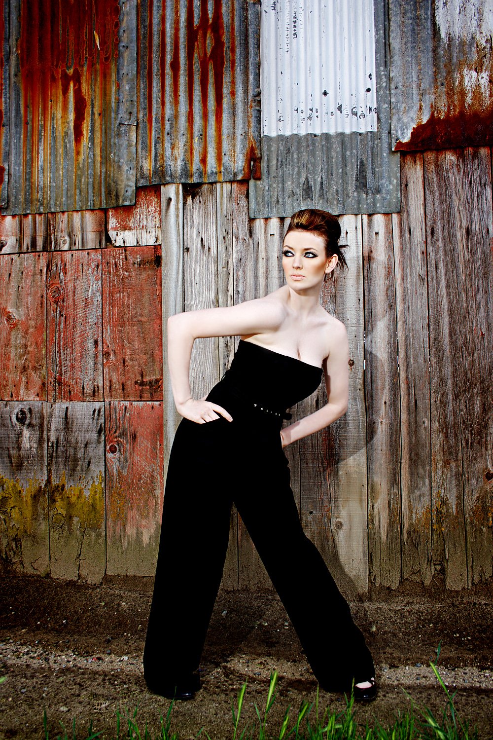 Woman posing against a weathered wooden wall in a black strapless wide-leg jumpsuit with a slim studded belt
