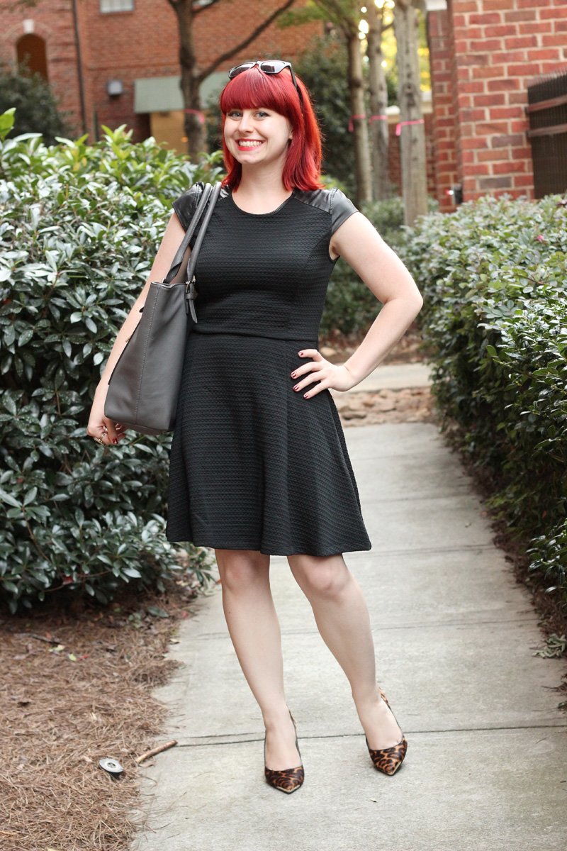 A woman with red hair wearing a black textured fit-and-flare dress with leather cap sleeves, carrying a gray work bag and wearing leopard-print heels, standing on a sidewalk