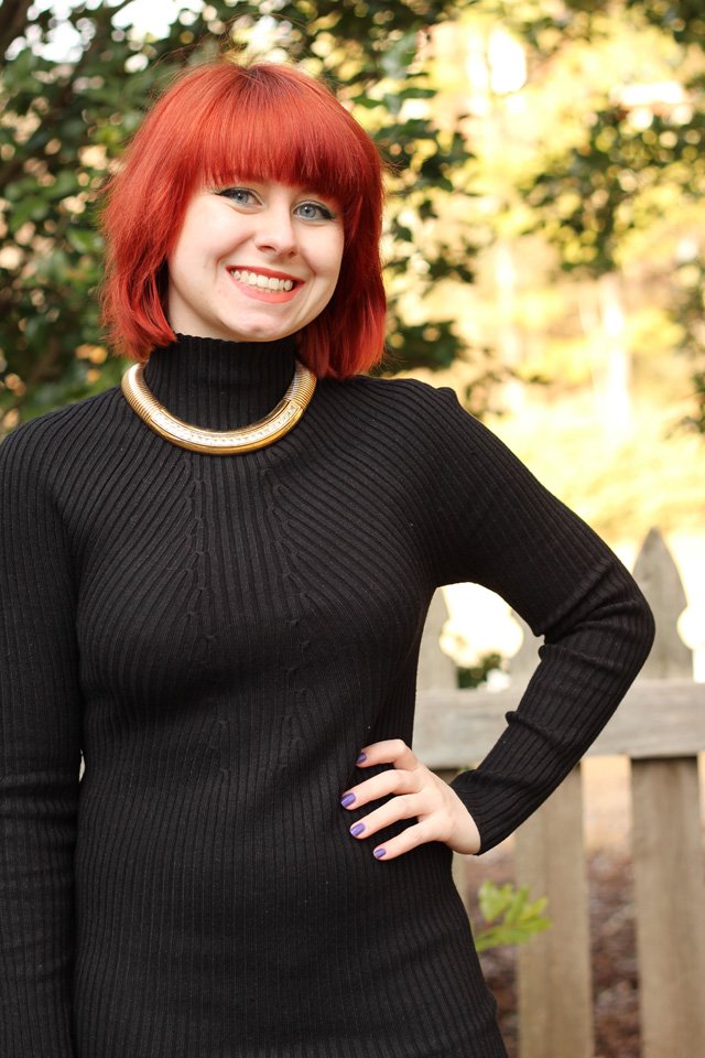A woman with a short red bob wearing a black ribbed turtleneck bodycon dress and a chunky gold choker necklace, smiling outdoors