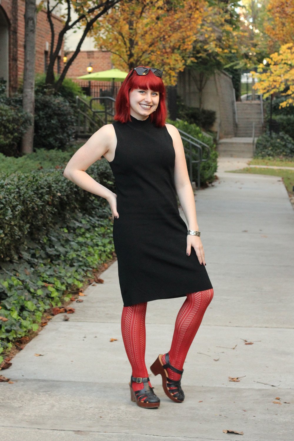 A woman with red bob hair wearing a black sleeveless turtleneck shift dress, bright orange patterned tights and black platform sandals, with sunglasses on her head