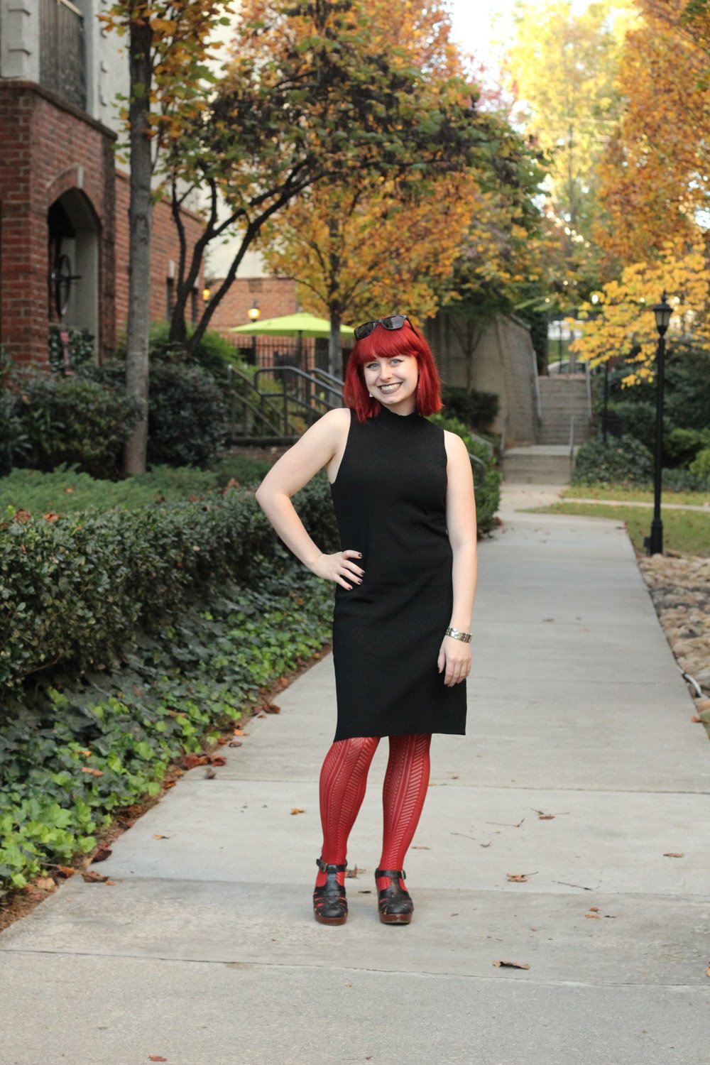 A woman with red hair wearing a black sleeveless turtleneck dress, rust-orange ribbed tights and dark heels, walking on a path lined with autumn trees