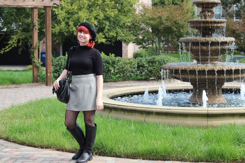 A woman with a red bob and black beret wearing a black short-sleeve turtleneck, gray houndstooth mini skirt, polka-dot tights and black knee-high boots, standing by a fountain
