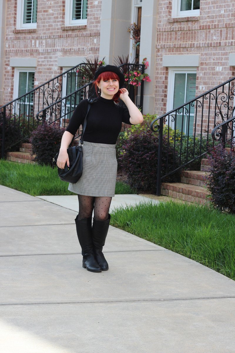 Smiling woman with red hair wearing a black beret, a black mock-turtleneck top, a grey houndstooth mini skirt, sheer polka-dot tights, and black knee-high boots, carrying a black shoulder bag, standing on a sidewalk in front of brick townhouses.