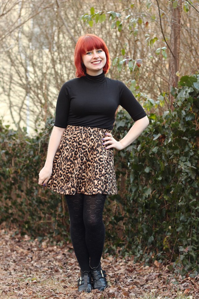 Smiling young woman with a bright red bob haircut standing outdoors among ivy bushes and fallen brown leaves, wearing a black long-sleeve turtleneck top with a clearly visible high neckline, a leopard-print flared skater skirt, black heart-pattern tights, and black strappy cutout ankle boots.