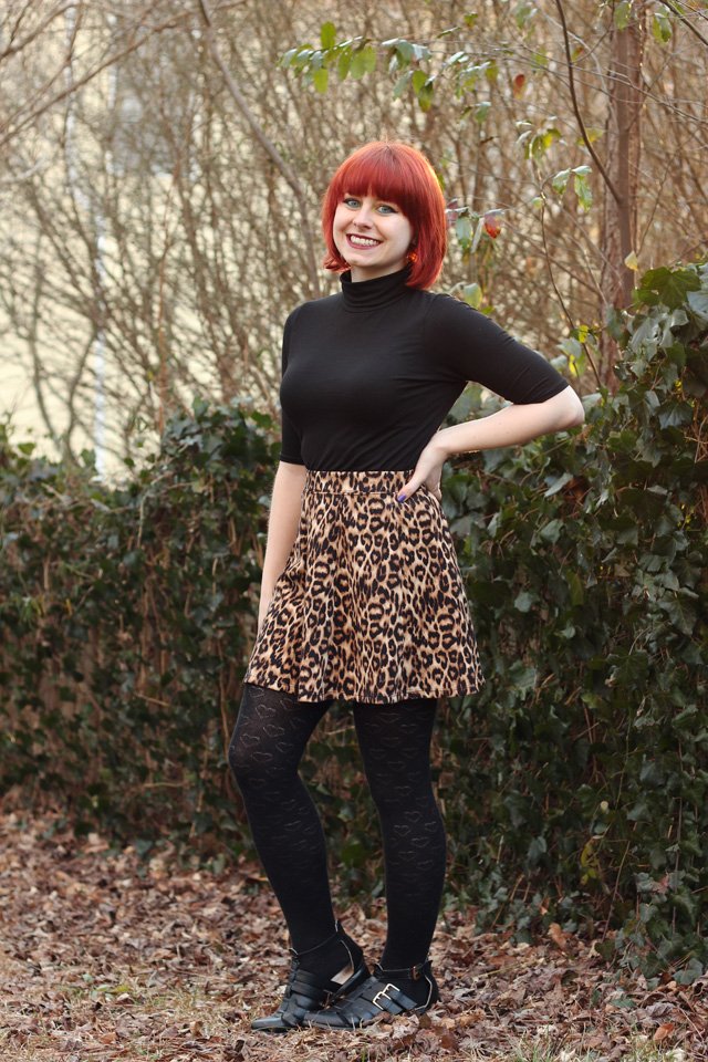A smiling red-haired woman outdoors wearing a black turtleneck top, a leopard-print skater skirt, black heart-print tights and black cutout ankle boots
