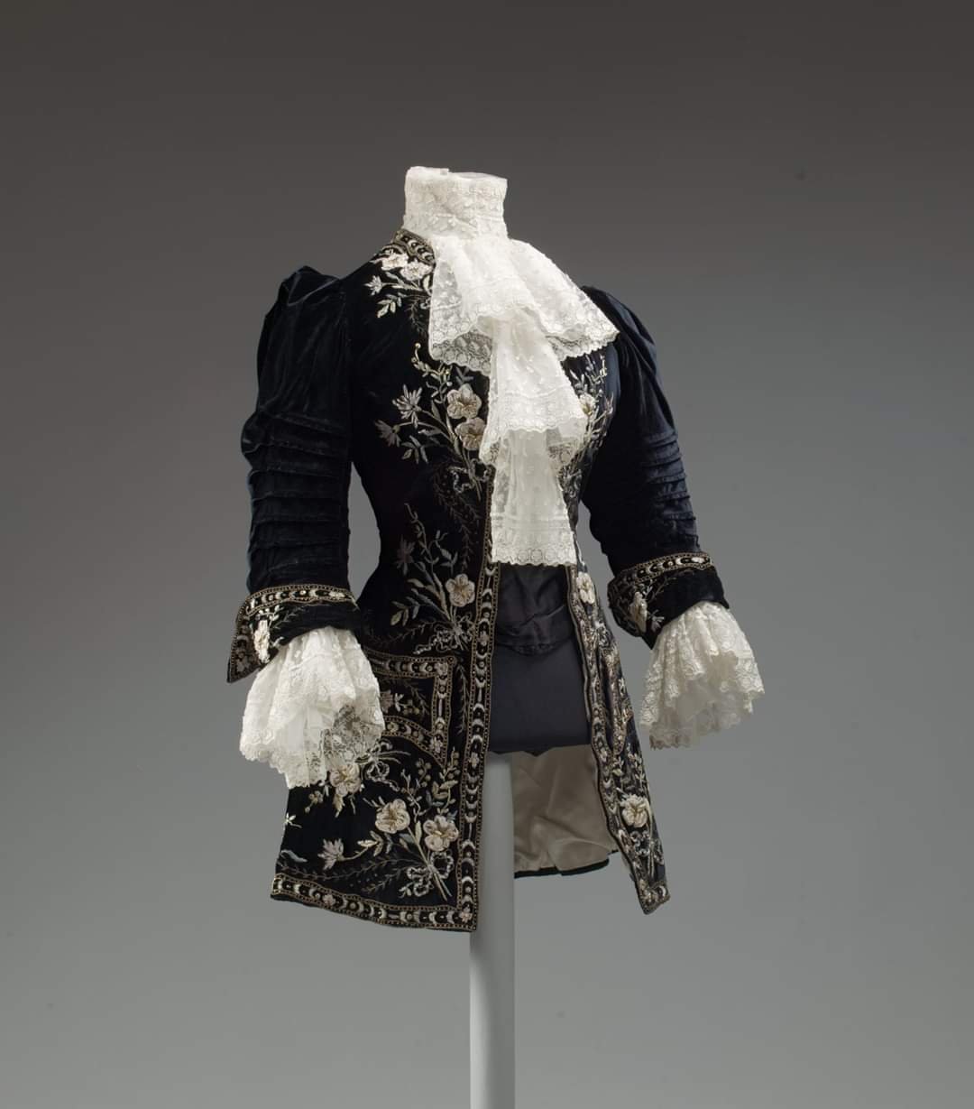 Color museum photograph of an 18th-century black velvet embroidered riding coat with lace stock and cuffs displayed on a mannequin