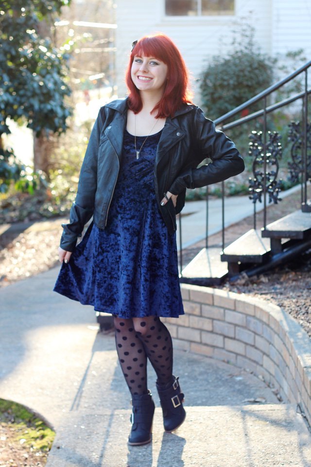 Red-haired woman outdoors wearing a blue crushed-velvet skater dress with a black leather jacket, polka-dot tights and black ankle boots.