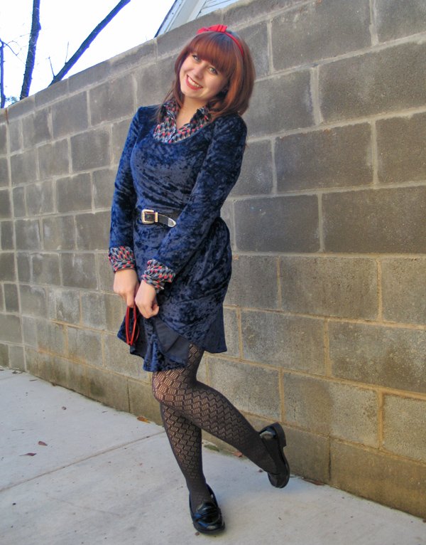Red-haired woman against a brick wall wearing a blue crushed-velvet long-sleeve dress with a patterned blouse collar and cuffs peeking out, a slim belt, patterned tights and black flats.