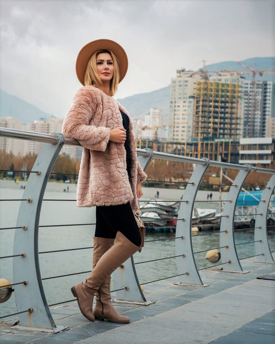 A blonde woman in a blush-pink faux-fur coat and camel fedora over a black dress with tan over-the-knee boots, leaning on a bridge railing by the water