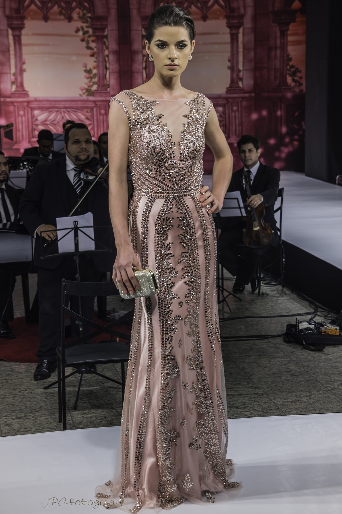 A model on a runway wearing a blush rose-gold embroidered full-length gown with a fitted bodice and an elegant silhouette.