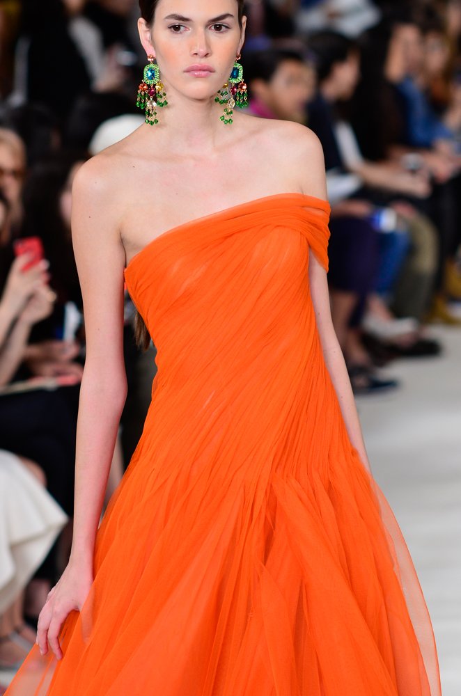 Model on a runway wearing a vivid tangerine-orange strapless pleated tulle gown with a draped neckline and statement chandelier earrings.
