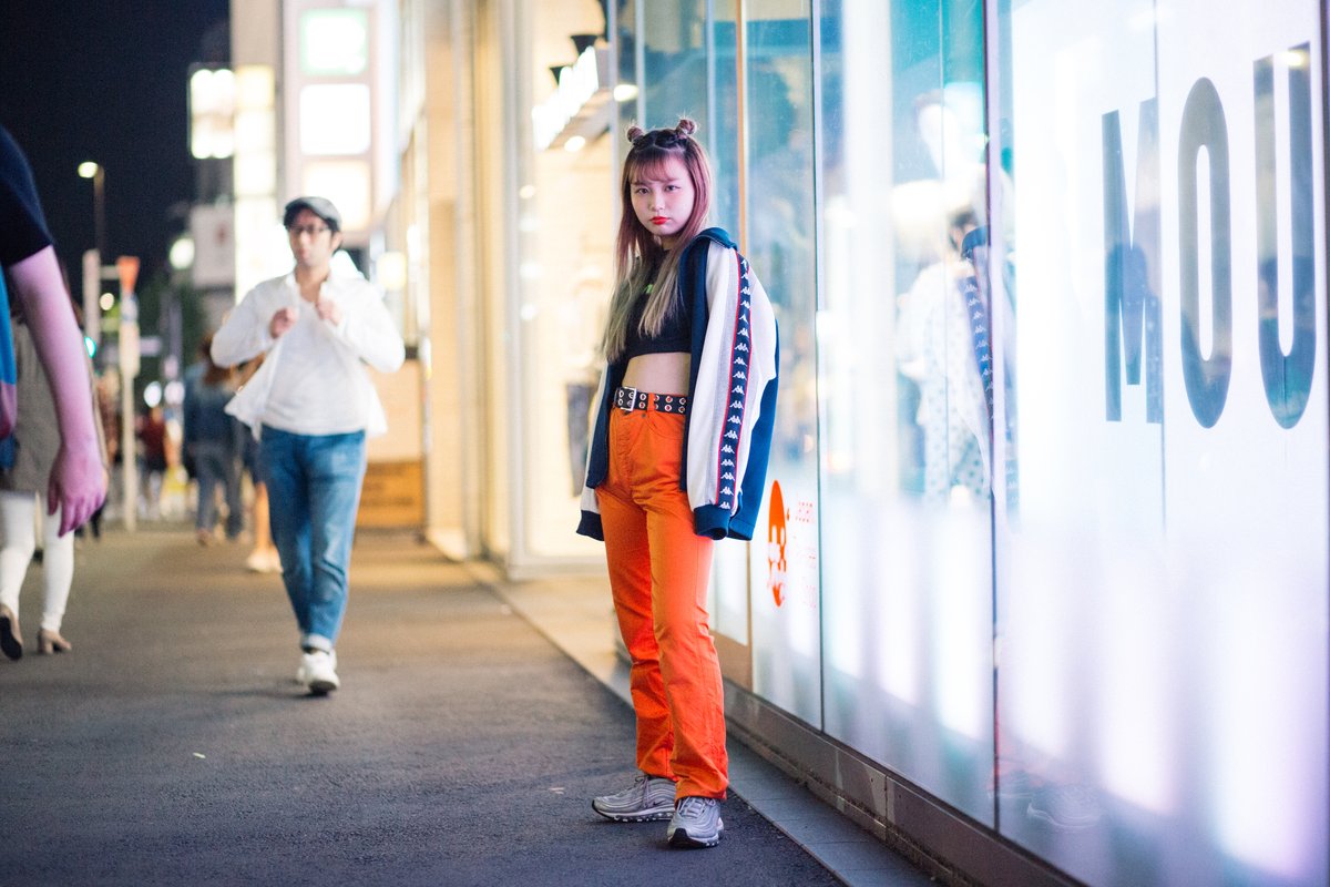 Young woman photographed at night in Harajuku wearing bright orange wide-leg trousers, a grommet belt, a black crop top and an open sporty jacket.