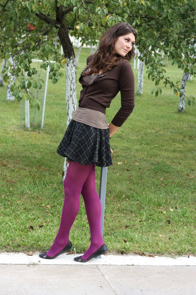 A woman outdoors wearing a brown fitted cardigan over a tan top with a black-and-green plaid pleated mini skirt, magenta opaque tights, and black ballet flats