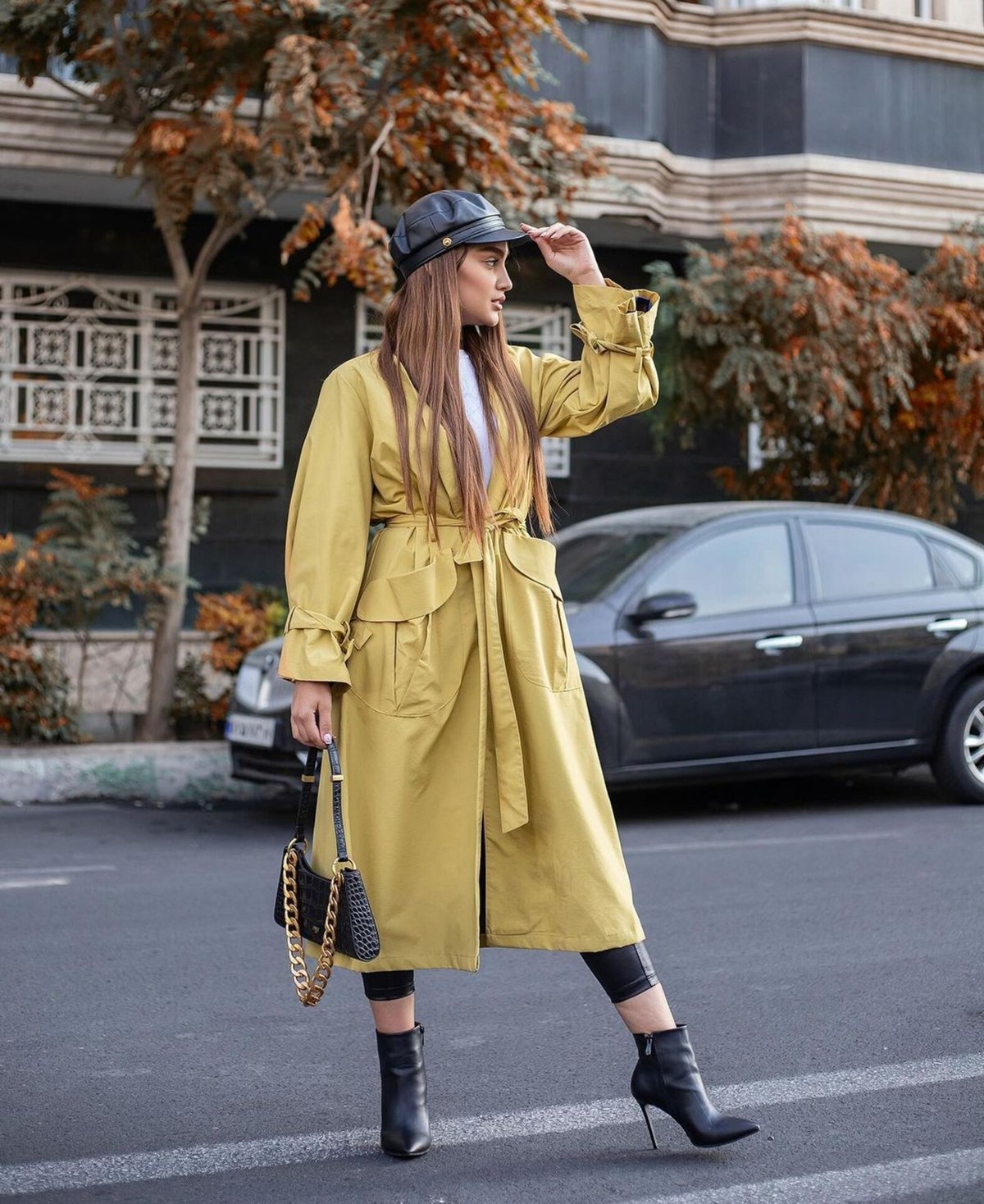 Woman in a pale butter-yellow belted trench coat with a black cap, black ankle boots and a black chain-strap handbag on a city street