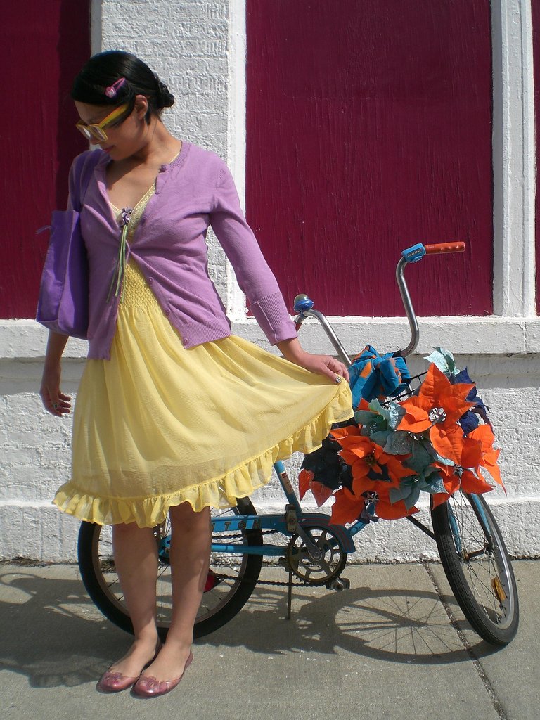 Woman in a pale yellow sundress with a purple knit cardigan, leaning on a flower-decorated bicycle