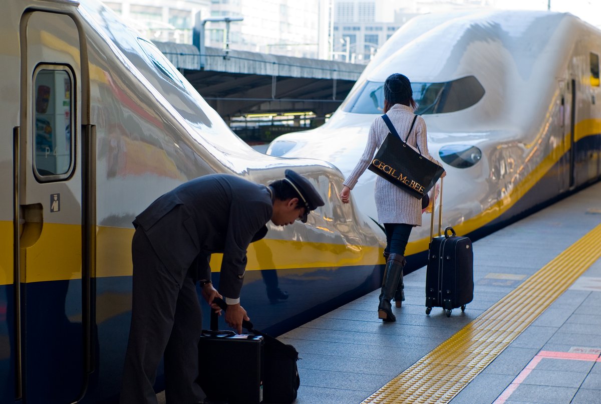A woman seen from behind on a Tokyo train platform wearing a light cable-knit sweater dress over dark tights with knee-high boots and a beanie, pulling a suitcase.