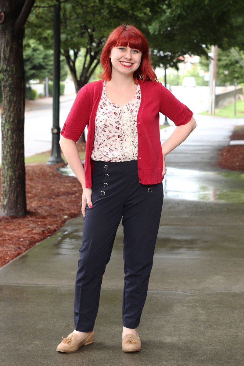 A woman with red hair wearing a red short-sleeve cardigan over a white floral blouse, navy sailor trousers and tan loafers, standing on a wet sidewalk