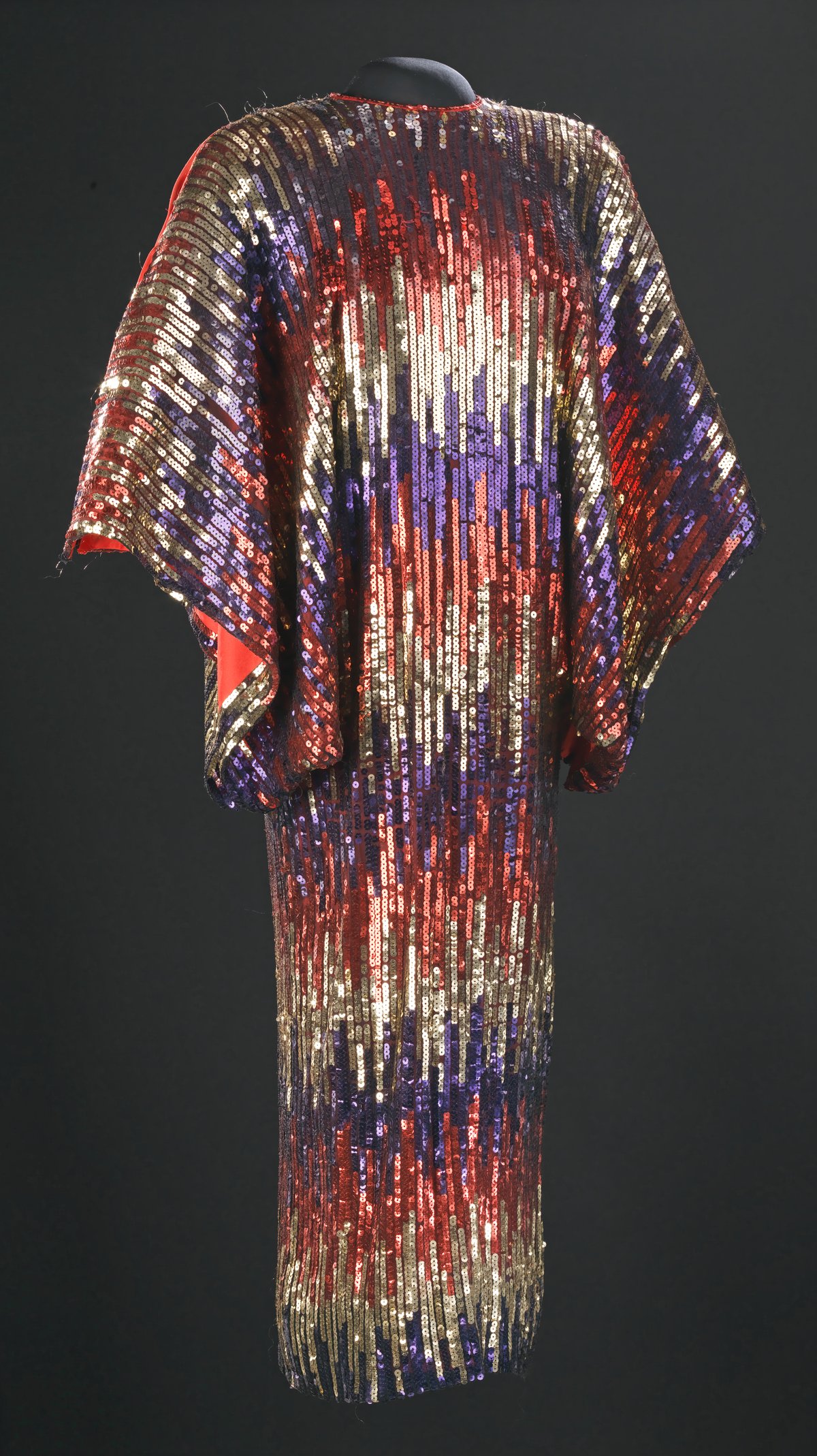A sequined caftan in gold, red and purple sequins displayed on a black mannequin against a dark background — Celia Cruz stage costume, Smithsonian collection