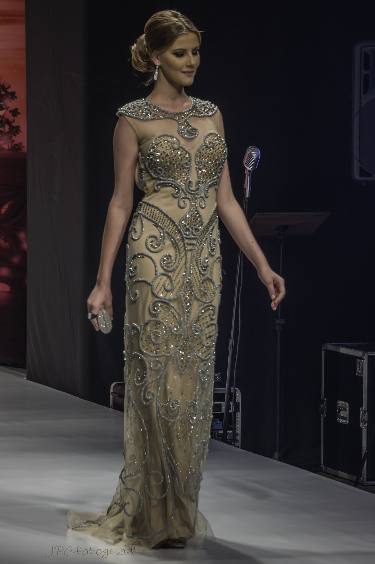 A model on a runway in a champagne-gold beaded full-length evening gown with a sheer embellished neckline and a flowing skirt.