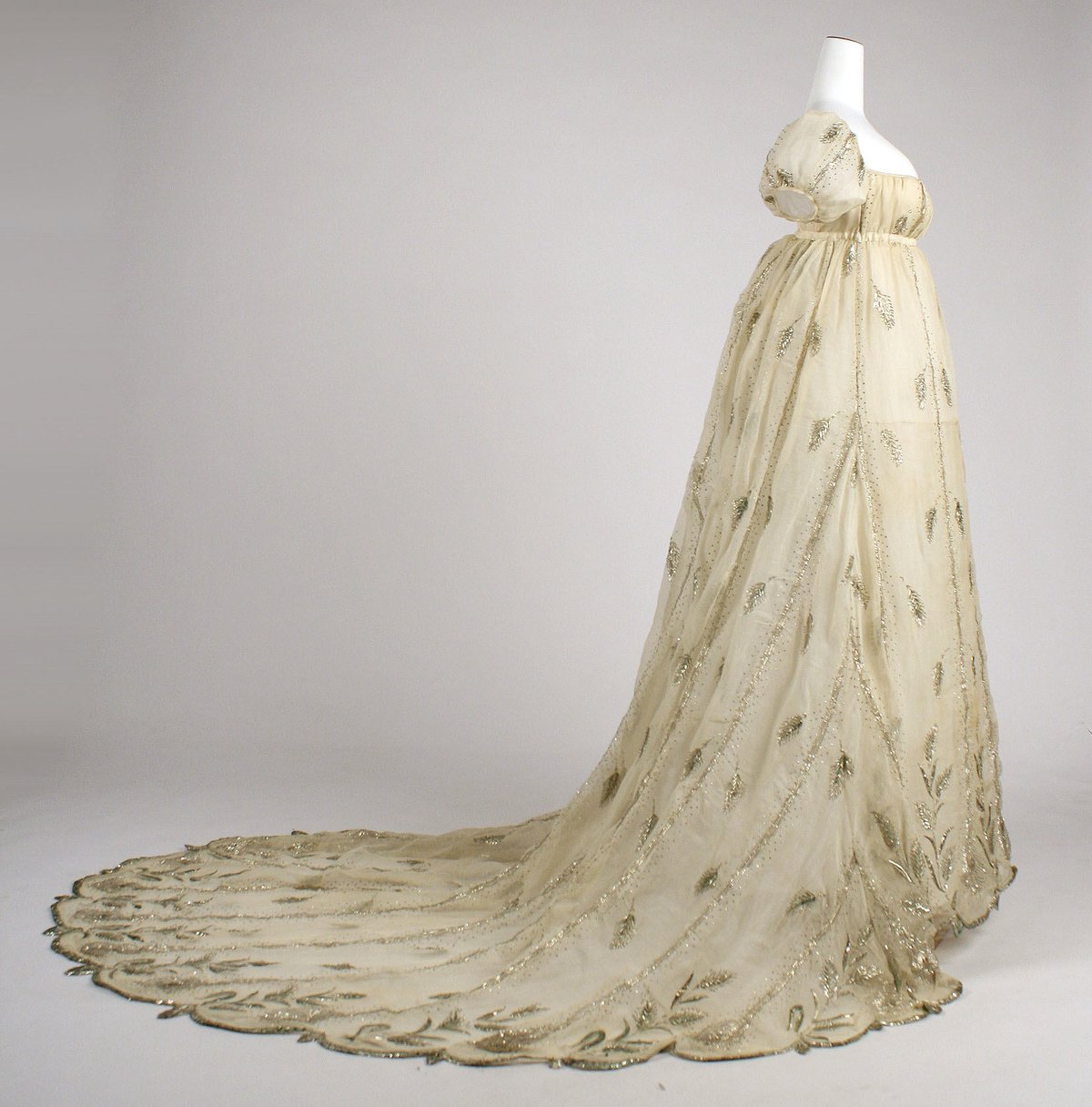 A champagne-colored empire-waist gown with gold metallic embroidery and a long train, displayed on a museum mannequin
