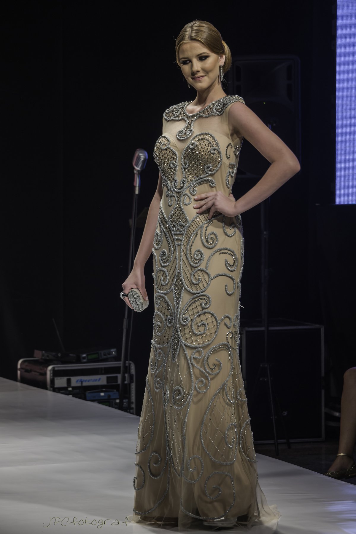 Model on a runway wearing a champagne evening gown with silver scroll beadwork and a sheer illusion neckline, holding a crystal clutch
