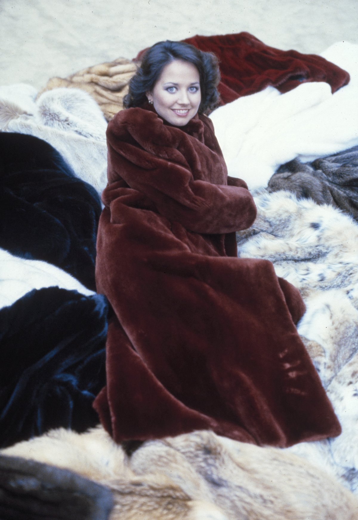 A smiling woman with dark curled hair in a rich chocolate-brown fur coat with a plush collar, seated among piles of fur coats.