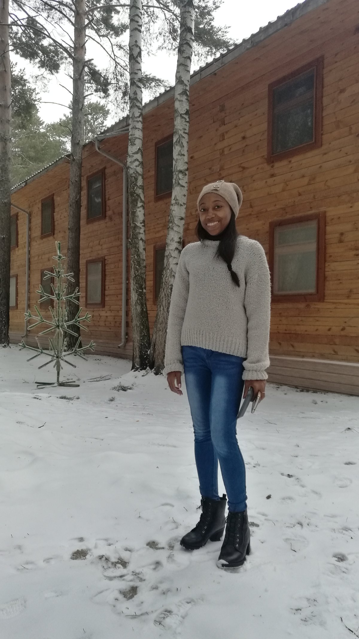 A smiling woman standing in snow beside a wooden cabin wearing a beige knit beanie, an oatmeal chunky knit sweater, blue skinny jeans and black lace-up winter boots