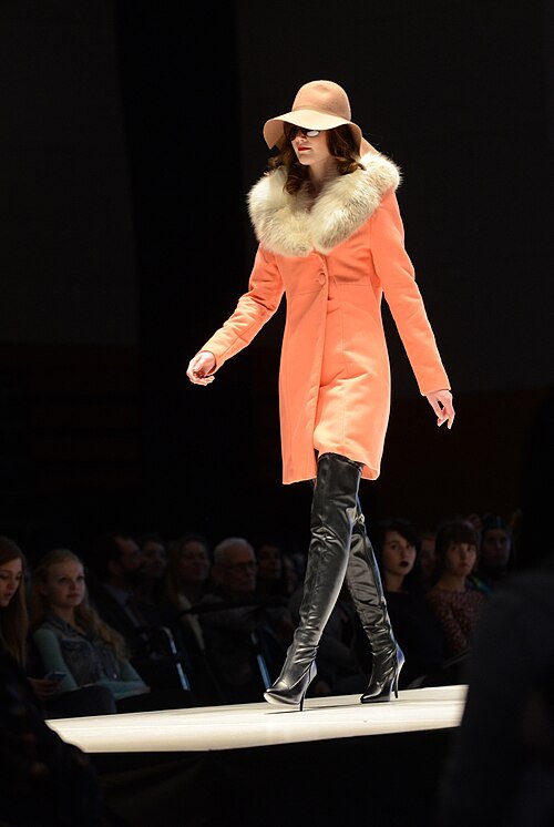 Model on a runway wearing a coral wool coat with a fur collar, black over-knee boots, and a wide-brim hat
