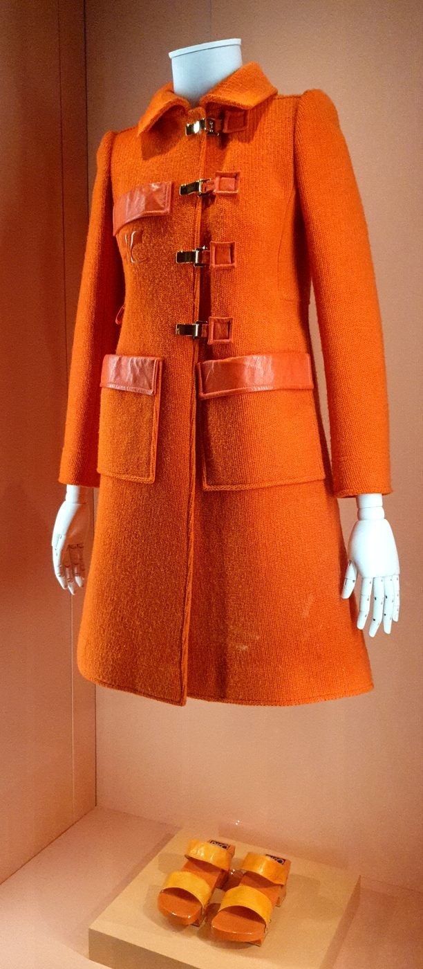 A 1972 Andre Courreges burnt-orange wool toggle coat with patch pockets and leather trim displayed on a museum mannequin, with matching orange sandals below