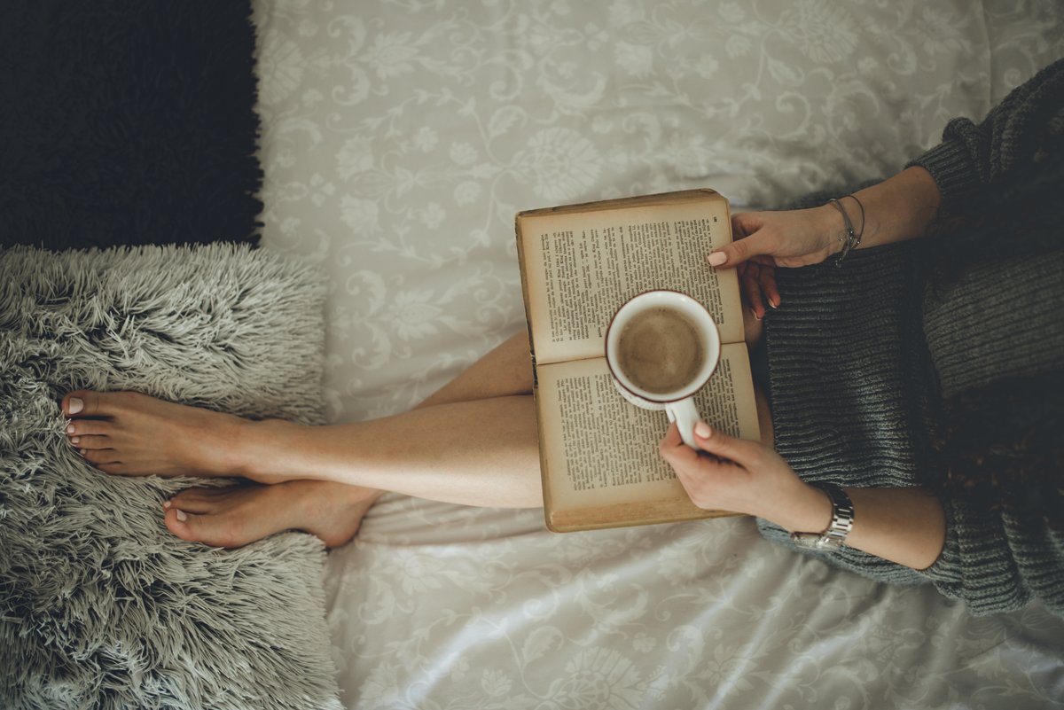 A woman lounging on a bed wearing an oversized chunky charcoal-grey knit sweater, holding an open book with a coffee mug
