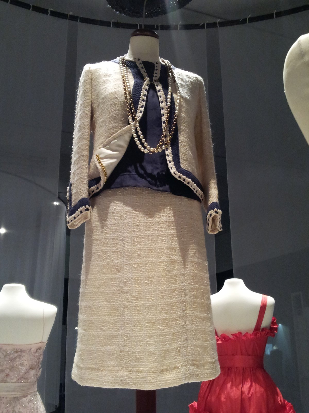 Museum mannequin displaying a late-1960s cream Chanel boucle tweed jacket and skirt with navy trim and layered pearl and chain necklaces.