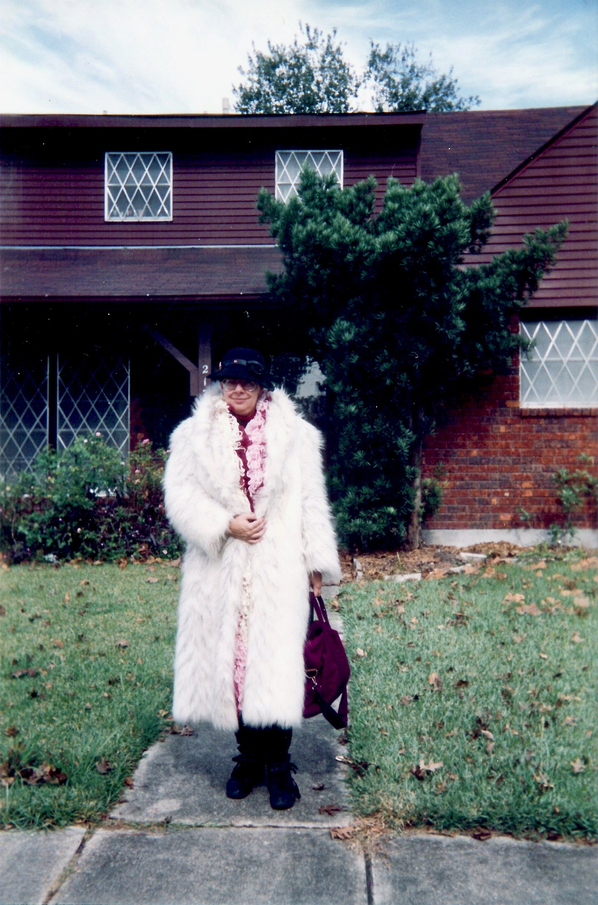 A woman on a suburban sidewalk in a long shaggy white faux fur coat worn open over a patterned dress, with dark tights, dark shoes and a burgundy handbag.