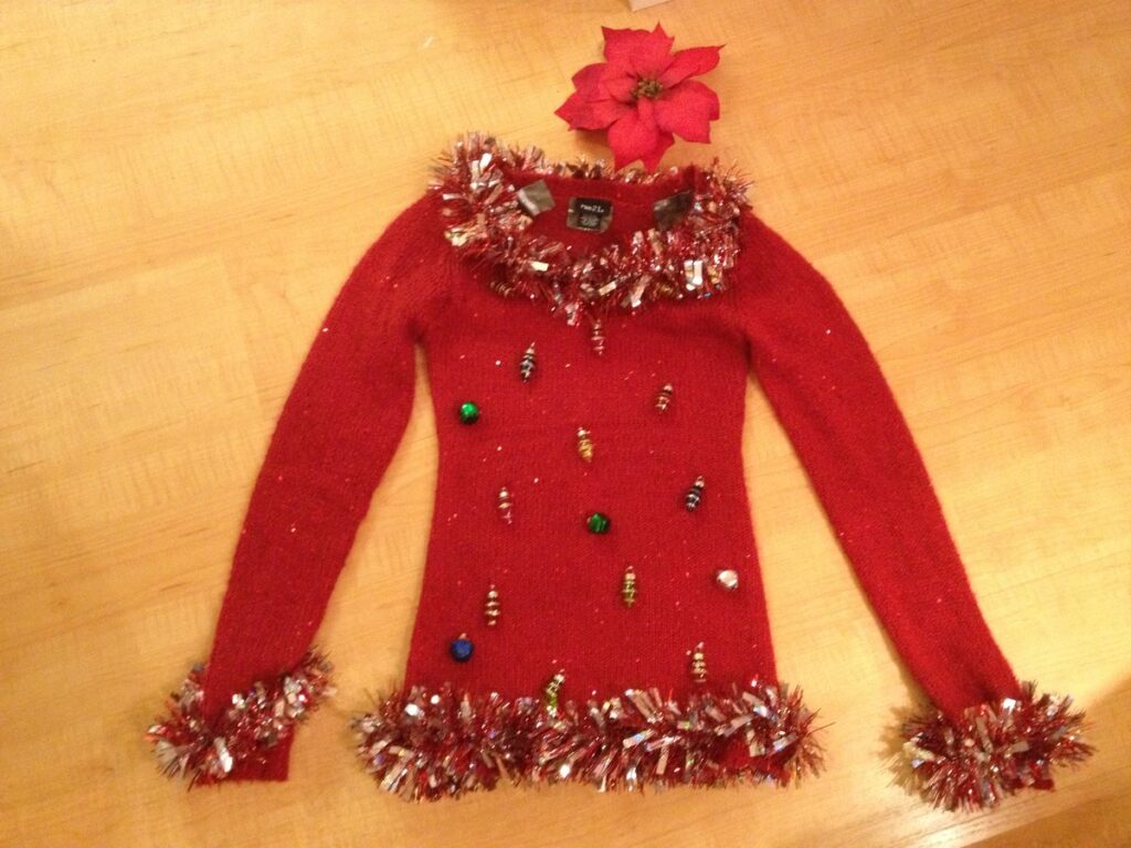 A red handmade ugly Christmas sweater decorated with tinsel trim, gold baubles and a red poinsettia laid flat