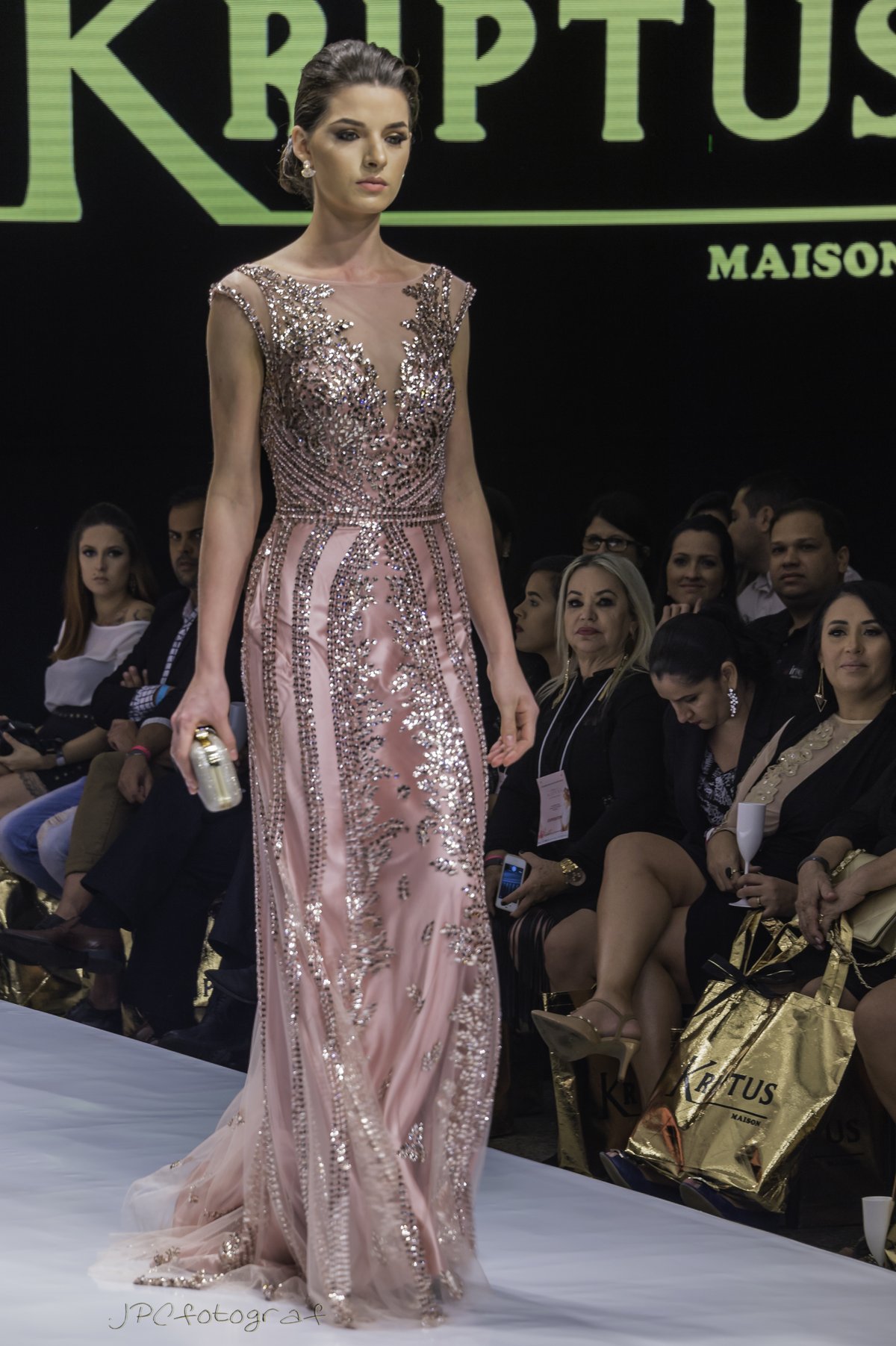 Model on a runway wearing a blush pink evening gown covered in silver sequin embellishment with an illusion neckline