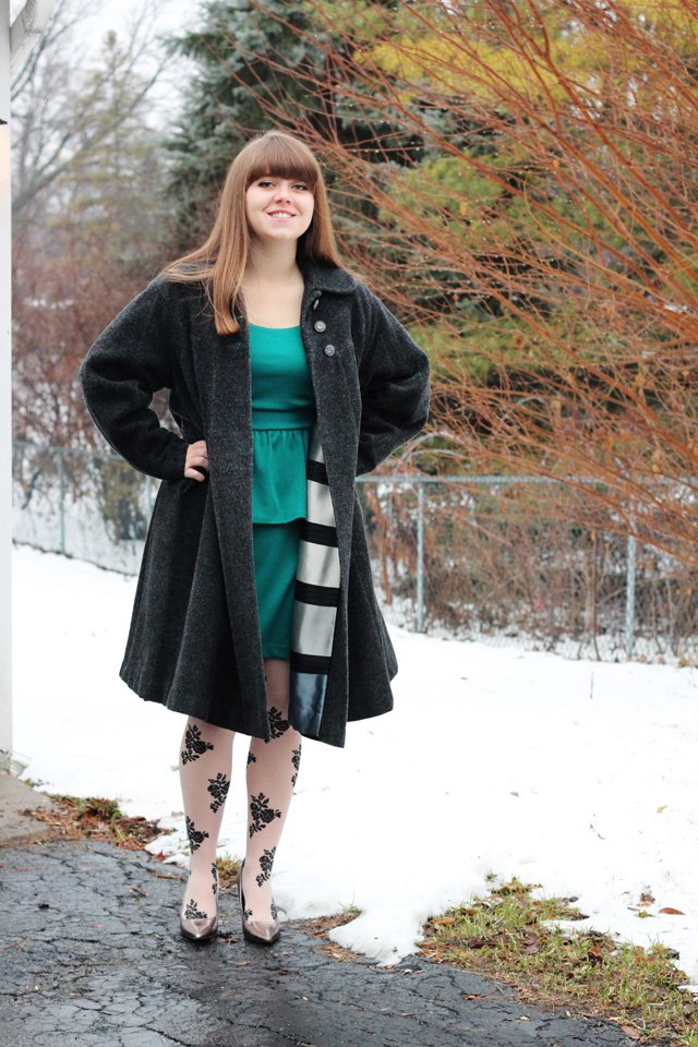 Smiling young woman standing in snow wearing a dark charcoal wool coat open over an emerald-green dress with floral tights and metallic heels
