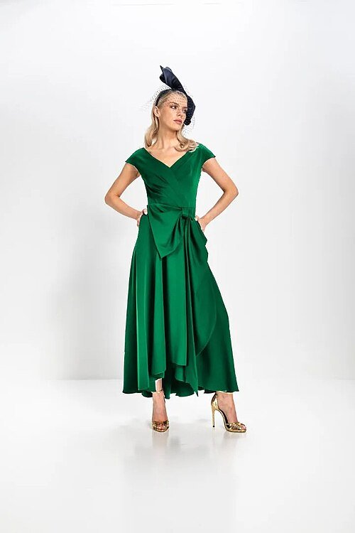 Model wearing an emerald green midi dress with a waist bow and a black headpiece, with gold heels