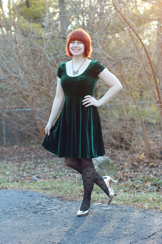 Red-haired woman outdoors wearing an emerald green velvet skater dress with bow-print tights and rose-gold heels.